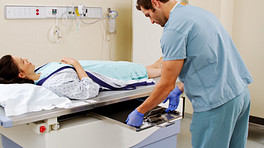 Patient lying on X-ray table while X-ray technician switches out the X-ray films beneath her.