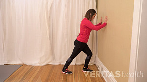 A woman is standing next to a wall, with her hands pressed against the wall. Her left leg is slightly bent with her toes almost touching the wall and her right leg parallel behind her to stretch her calf.