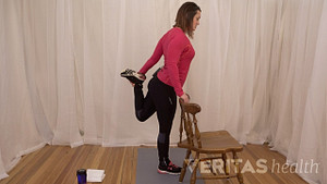 Woman standing next to a chair, and holding onto it with one hand for support, with the other hand she grabs her heel from behind her and pulls it towards her buttocks to stretch the quadricep muscle.