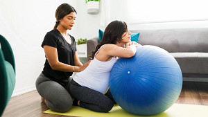 Pregnant woman leaning on exercise ball with trainer helping stretch the lower back.